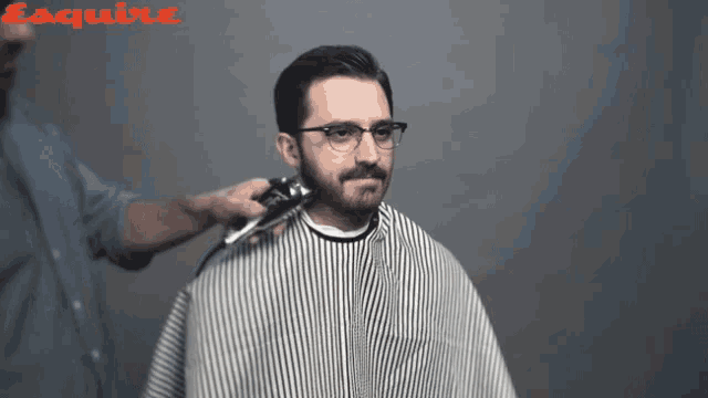 a man with glasses is getting his hair cut by a man wearing a cape
