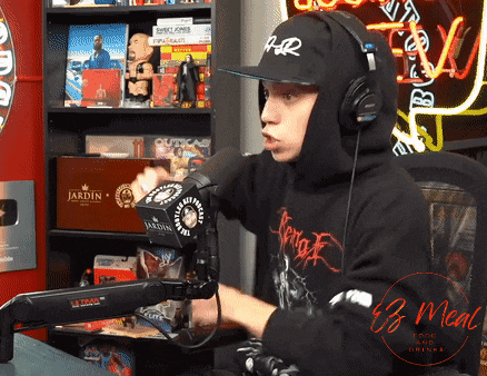 a man wearing headphones and a black hoodie is talking into a microphone in front of a sign that says jardin