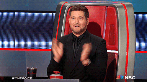 a man in a suit applauds while sitting in a chair with a nbc logo behind him