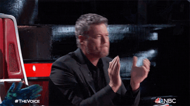 a man applauds while sitting in front of a chair that says nbc on it