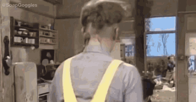 a man in a gray shirt and yellow overalls is standing in a kitchen with his back to the camera .