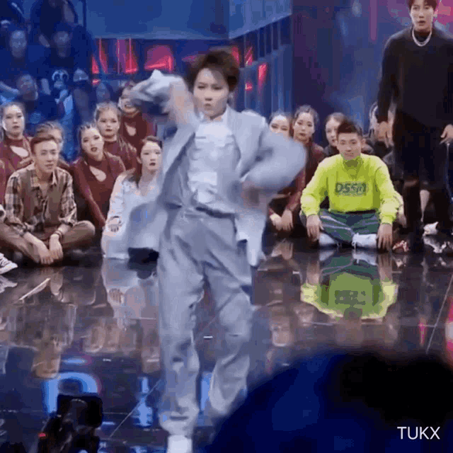 a group of people are sitting on the floor watching a man in a grey suit dance on a stage .