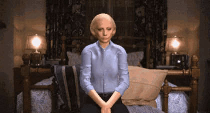 a woman in a blue shirt sits on a bed with her hands folded