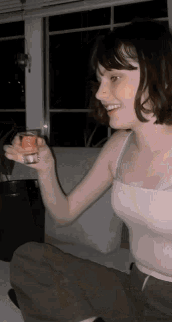 a woman in a white tank top is smiling and holding a shot glass