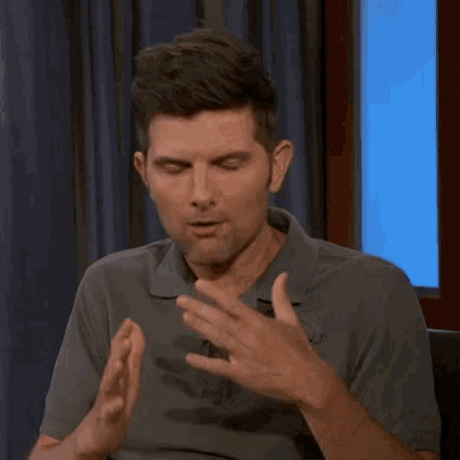 a man in a gray shirt is sitting in front of a blue curtain and making a funny face with his hands .