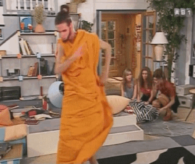 a man in an orange robe is dancing in a living room while a group of people watch .