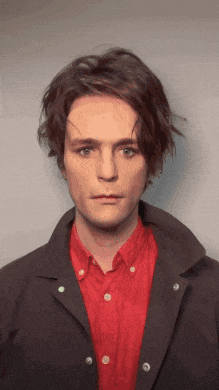 a young man wearing a red shirt and a black jacket looks at the camera
