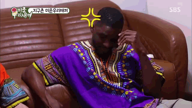 a man in a purple shirt is sitting on a couch with a sbs logo behind him