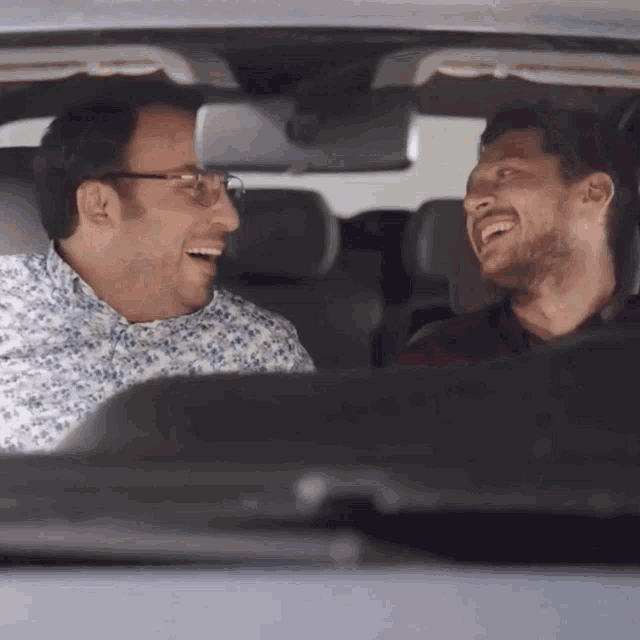 two men are laughing in a car and one of them is wearing glasses