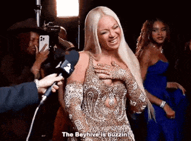 a woman in a gold dress is being interviewed by a reporter while holding her chest .