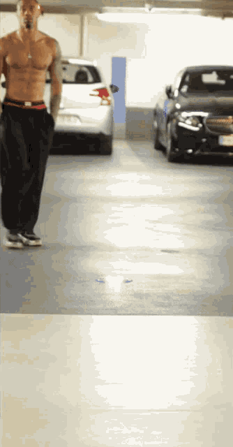 a shirtless man walks through a parking garage with cars parked behind him
