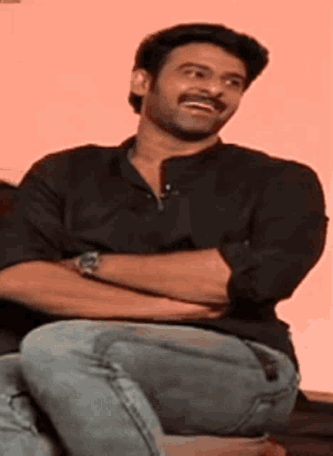 a man wearing a black shirt and jeans is sitting with his arms crossed and smiling .