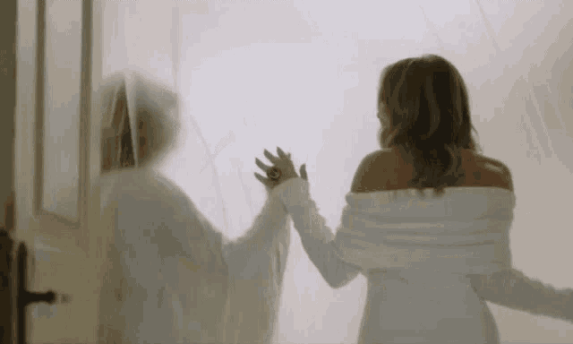 a woman in a white robe is standing in front of a door with a ghost in the background .