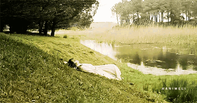 a woman in a white dress is laying in the grass next to a body of water