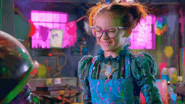 a little girl wearing glasses and an apron is smiling in a room .