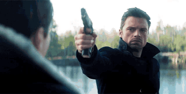 a man is pointing a gun at another man while standing next to a body of water .
