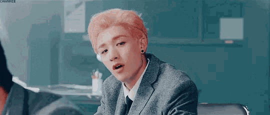 a young man with pink hair is wearing a suit and tie and sitting in a chair .
