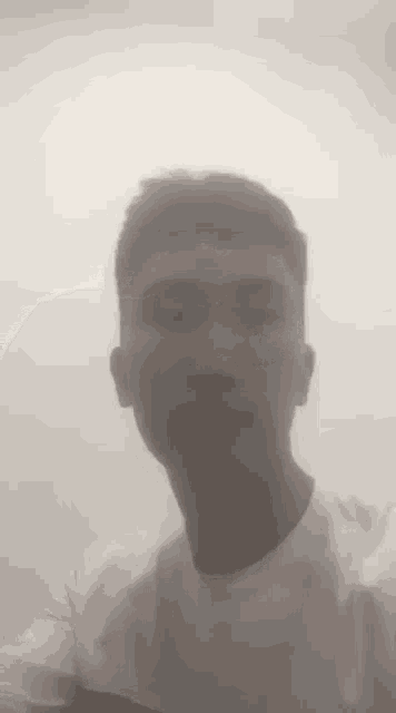 a man in a white t-shirt is taking a selfie in front of a foggy wall .