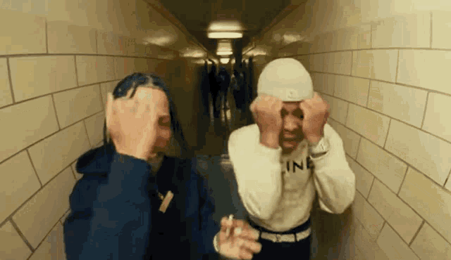 two men are standing next to each other in a hallway covering their faces with their hands .