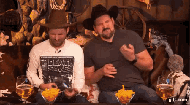 two men wearing cowboy hats are sitting at a table with drinks and snacks .
