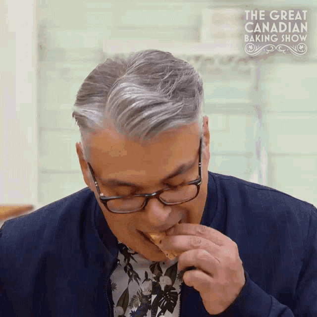 a man wearing glasses is eating a piece of food with the great canadian baking show in the background