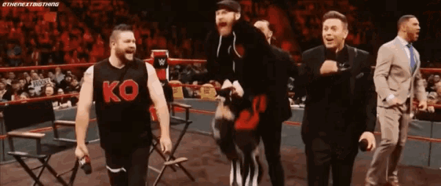 a group of men are standing in a wrestling ring and one of them is wearing a ko shirt .