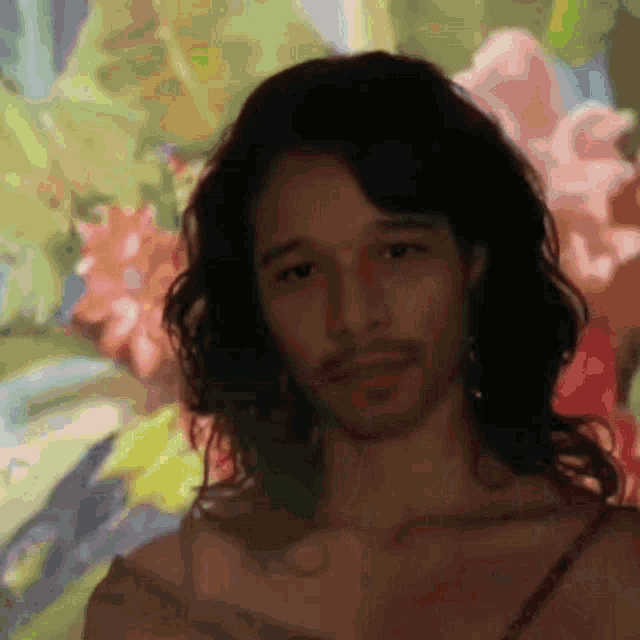 a man with long hair and a beard stands in front of flowers