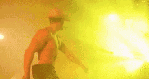 a shirtless man wearing a cowboy hat is dancing in front of a yellow light .