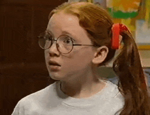 a young girl with red hair and glasses is wearing a white shirt and a red bow in her hair .