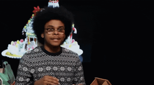a man wearing glasses and a sweater with snowflakes on it stands in front of a gingerbread house