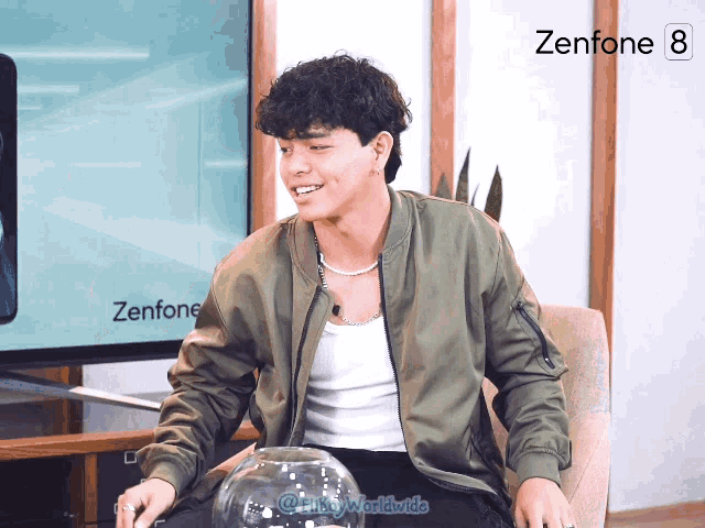 a man in a green jacket is sitting in front of a zenfone 8 screen