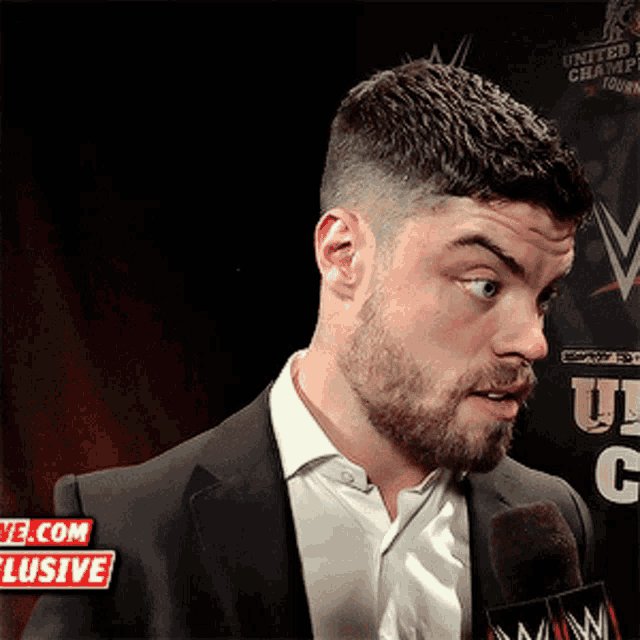 a man in a suit is talking into a microphone in front of a sign that says ' wwe.com exclusive '