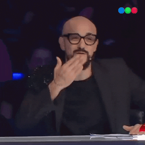 a bald man wearing glasses and a black jacket covering his mouth with his hand