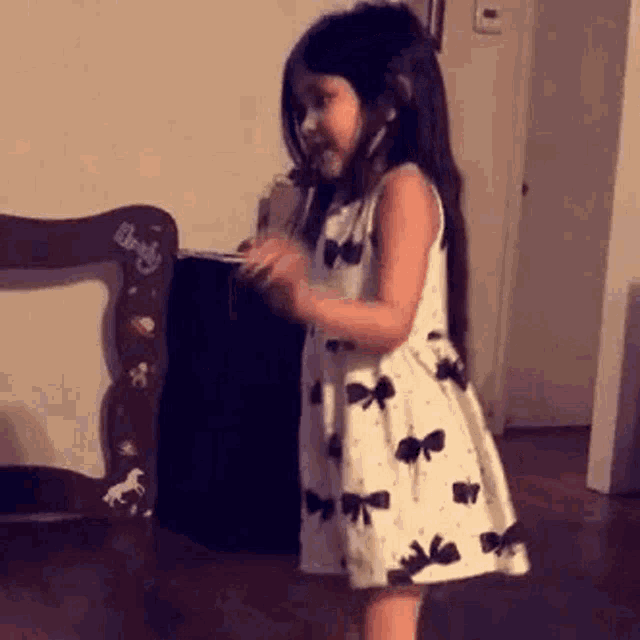 a little girl in a white dress is standing in front of a wooden chair in a room .