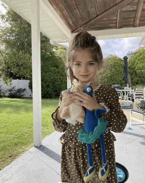 a little girl in a brown polka dot dress holds a stuffed animal