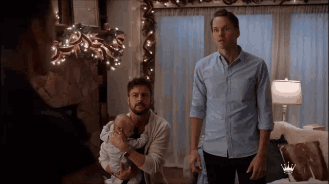 two men holding a baby in a living room with a crown on their shirt
