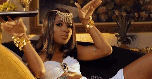 a woman in a white top and gold jewelry is sitting on a couch with her hands in the air
