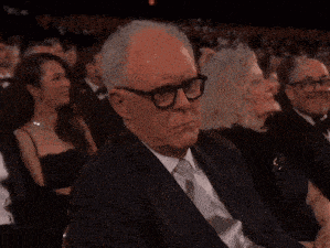 a man wearing glasses and a suit sits in a crowd of people clapping