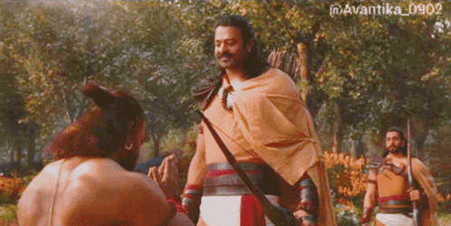 a man with a bow and arrow talks to another man in a forest
