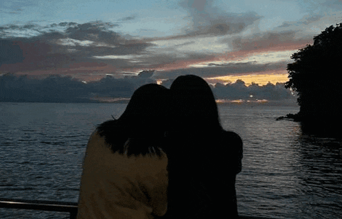 two women are hugging each other in front of a body of water at sunset .