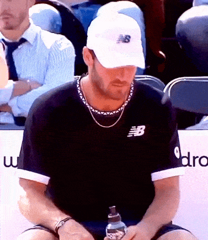 a man wearing a new balance hat sits in a stadium holding a bottle of water