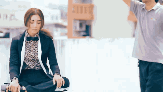 a woman in a suit sits in a lotus position while a man in a purple shirt stands behind her