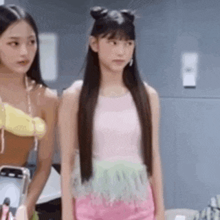 two young women are standing next to each other in a room . one of the girls is wearing a pink top and pink shorts .