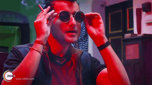 a man wearing round sunglasses is smoking a cigarette in a room with a zee5 logo behind him