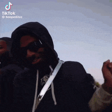 a man wearing a hoodie and sunglasses is standing in front of a group of people .