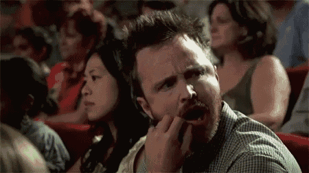 a man with a beard is sitting in front of a crowd of people in a theater and making a funny face .