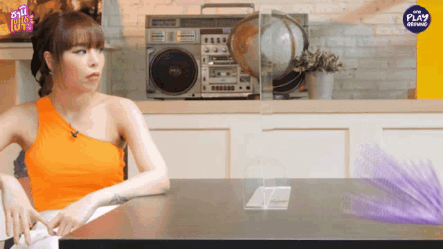 a woman in an orange top sits at a table in front of a boombox that says the play on it