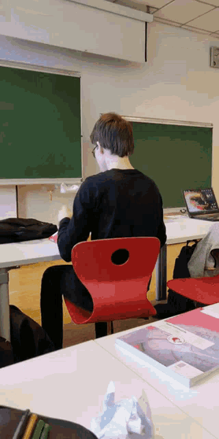 a boy sits in a red chair in front of a blackboard with a laptop on it