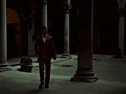 a man in a brown suit and white shirt stands in a dark courtyard