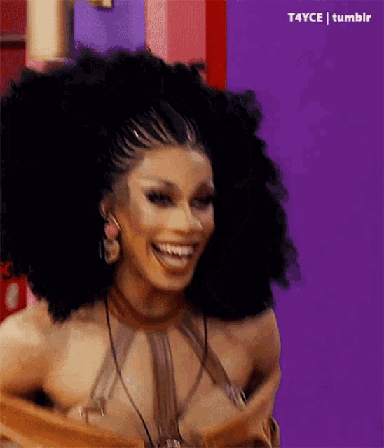 a drag queen with a braided ponytail and a big afro is smiling on a purple background .